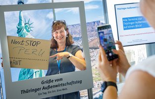 Eine Frau fotografiert eine andere Frau, die hinter einem weißem Bilderrahme mit der Aufschrift: Grüße vom Thüringer Außenwirtschaftstg, steht. Die fotografierte Frau hält dabei ein Schild hoch auf dem: Next Stop - Peru, zu sehen ist.