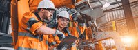 Zwei Arbeiter in orangefarbener Schutzausrüstung schauen in einer Fabrik auf ein Tablet.