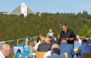 Teilnehmer der Veranstaltung essen gemeinsam auf der Terrasse des Veranstaltungsorts. Im Hintergrund ist das AHORN Panorama Hotel in Oberhof.