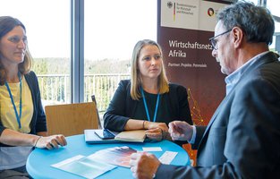 Zwei Frauen unterhalten sich mit einem Mann über das Unternehmen, welches sie vertreten. Sie stehen dabei an einem Stehtisch.