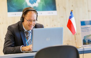 Ein Mann nimmt am Laptop an einer Video-Konferenz teil. Neben ihm steht eine kleine Flagge des Landes Chile.