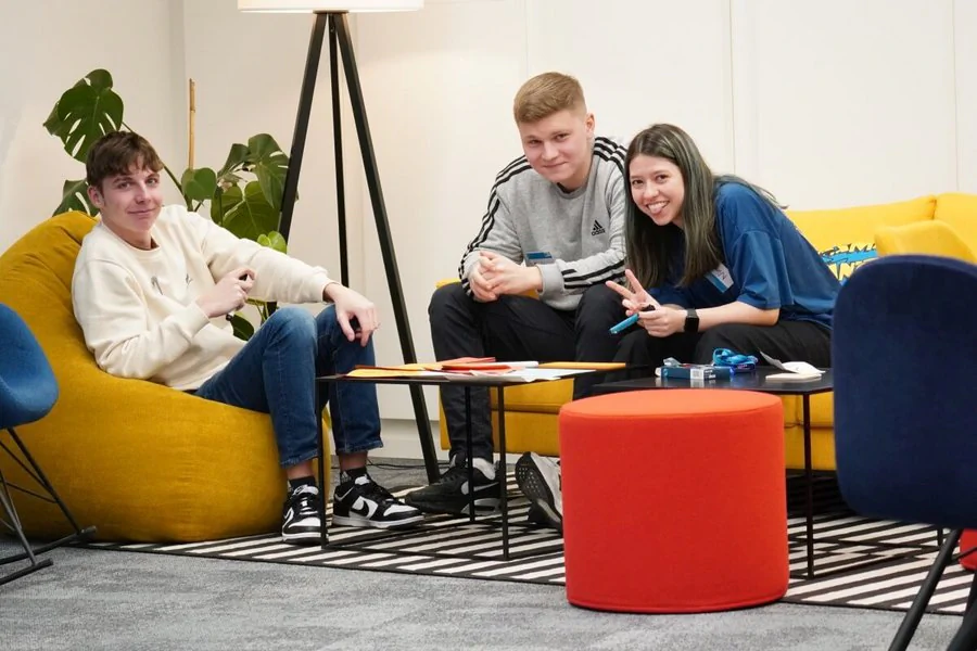 Drei Personen sitzen auf einem gelben Sessel und einer Couch in einem modernen Meetingraum