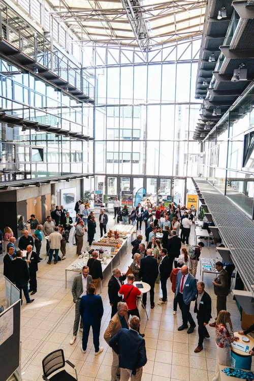 Copyright: TMDI / B. Schmidt Large event in a light-flooded glass atrium: Numerous people stand at high tables and information booths, chat, and use the catering offer in the middle of the hall.