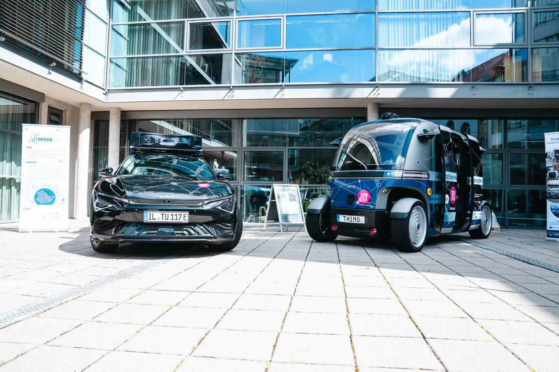 Copyright: TMDI / B. Schmidt Autonomous shuttle vehicle and black test vehicle with roof sensors are parked in front of a modern office building with a glass facade.