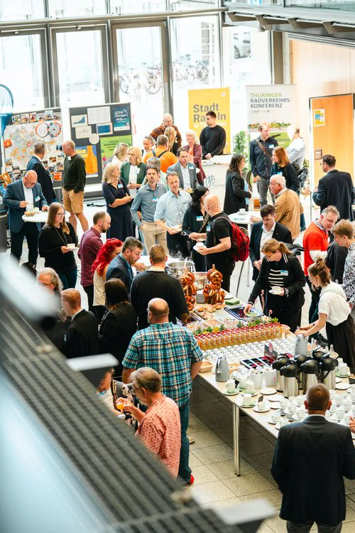 Copyright: TMDI / B. Schmidt Large networking event in a bright foyer: Numerous participants stand together in groups, chat at standing tables, and use a catering buffet with drinks and snacks.