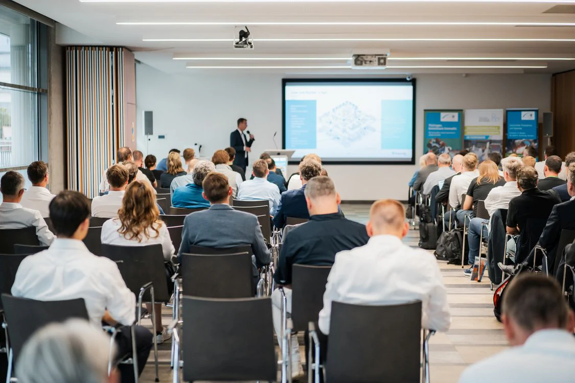 Publikum sitzt in einem Konferenzraum und verfolgt einen Vortrag mit Präsentation auf einer großen Leinwand.