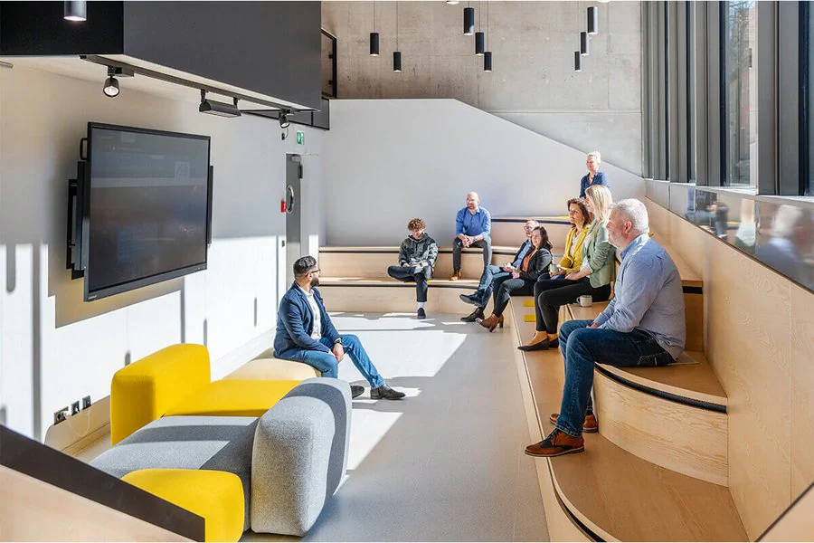 Menschen sitzen auf Bänken in einem modernen Meetingraum, umgeben von zeitgemäßer Architektur und ansprechendem Design.
