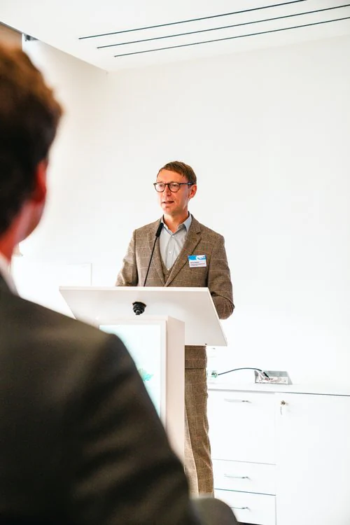 Copyright: TMDI / B. Schmidt Person stands at a lectern with a microphone and gives a presentation in a bright conference room. In the foreground, a listening person is visible out of focus.