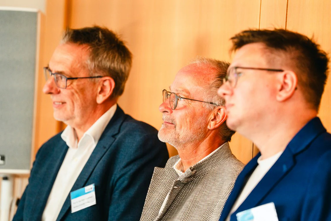 Copyright: TMDI / B. Schmidt Three people in business attire sit side by side at an event and attentively watch a presentation outside the picture.