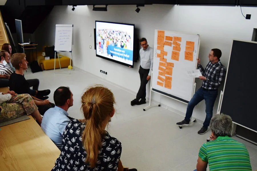 Eine Gruppe von Personen sitzt in einem Raum, während sie an einem Whiteboard diskutieren und Ideen austauschen.