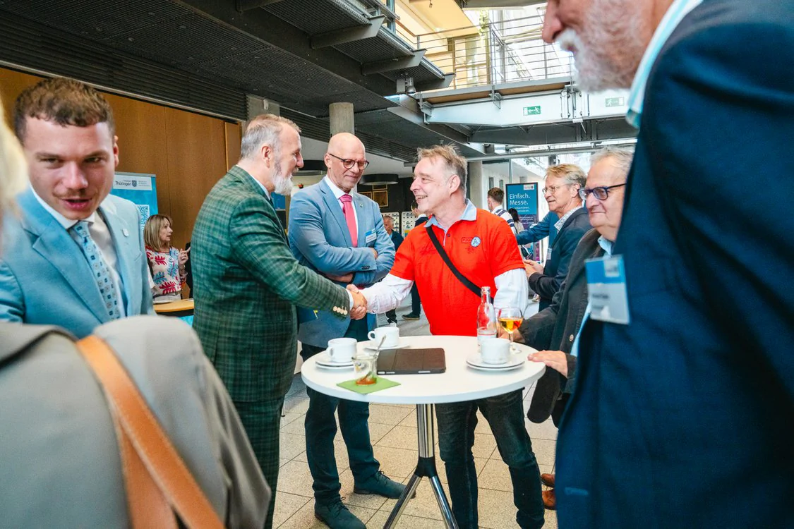 Copyright: TMDI / B. Schmidt Several participants of a network or industry event meeting stand around a high table in a modern foyer and engage in conversations. Two people greet each other with a handshake, while other guests in the background talk to each other at information booths and tables.