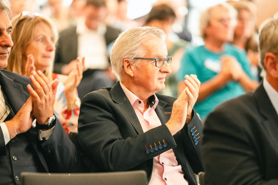 Copyright: TMDI / B. Schmidt Several seated people at an event are applauding. In the foreground, there is a person with glasses in a dark jacket; in the background, more participants are visible, depicted out of focus.