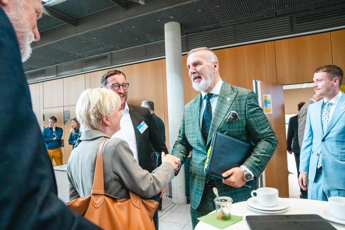 Copyright: TMDI / B. Schmidt People greet each other with a handshake at a networking event in a modern conference area; on a standing table in the foreground are coffee cups and a glass of drink.