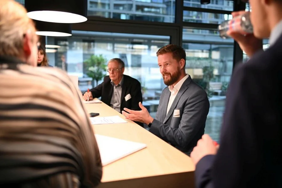 Ein Mann in einem Anzug führt ein Gespräch mit mehreren Personen, die an einem Tisch sitzen, und beteiligt sich aktiv am Austausch.