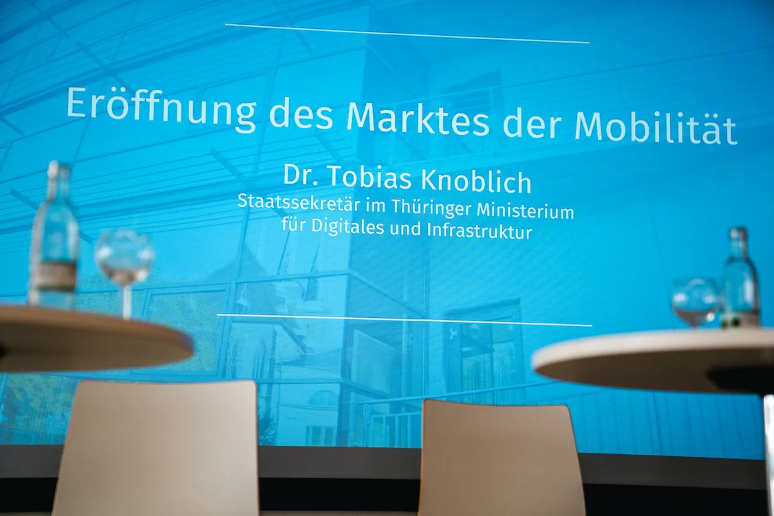 Copyright: TMDI / B. Schmidt Presentation slide on an event stage with the title “Opening of the Mobility Market“. In the foreground, there are two standing tables with water carafes and glasses, as well as two chairs. On the screen, “Dr. Tobias Knoblich, State Secretary at the Thuringian Ministry for Digital and Infrastructure“ is mentioned.