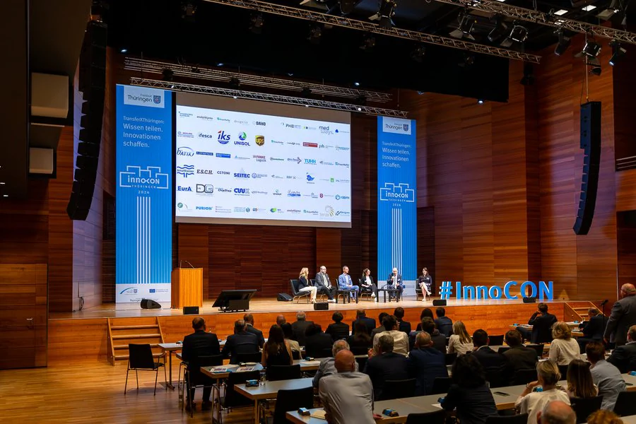 InnoCON Bildergalerie Foto: Menschen sitzen an Tischen in einem großen Auditorium und schauen der Podiumsdiskussion zu