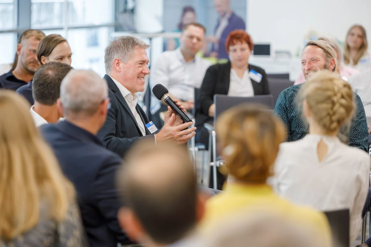Angebote für Netzwerkpartner | Bild: LEG Thüringen Bildbeschreibung / description: Mehrere Personen sitzen in einem modernen Veranstaltungsraum im Kreis und verfolgen einen Beitrag mit Mikrofon – Szene für Austausch, Dialog und Angebote für Netzwerkpartner.