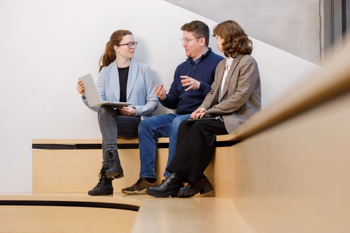 Bildbeschreibung / description: Drei Personen sitzen auf einer Holzstufe in einem modernen Innenraum und unterhalten sich miteinander. Eine Person hält ein aufgeklapptes Laptop auf dem Schoß, eine andere gestikuliert mit den Händen, während die dritte Person zuhört.