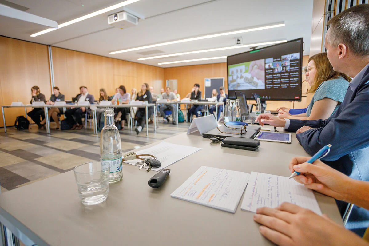 Bildbeschreibung / description: Teilnehmer einer Konferenz halten hybrid eine Besprechung ab