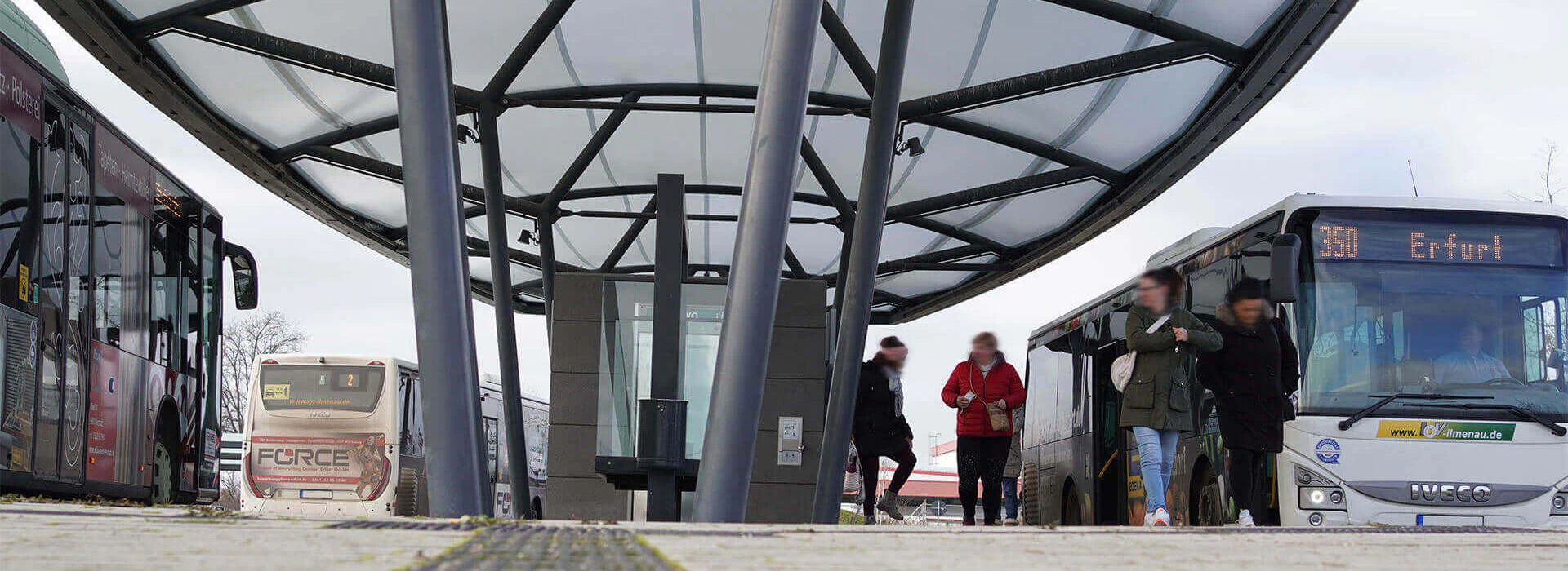 Image description: People get on and off a bus at a bus stop, while others wait on the street